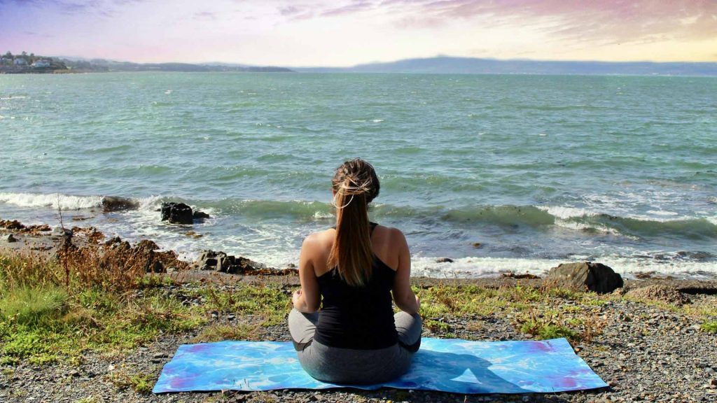 A person sits on a blue yoga mat, overlooking a coastline.