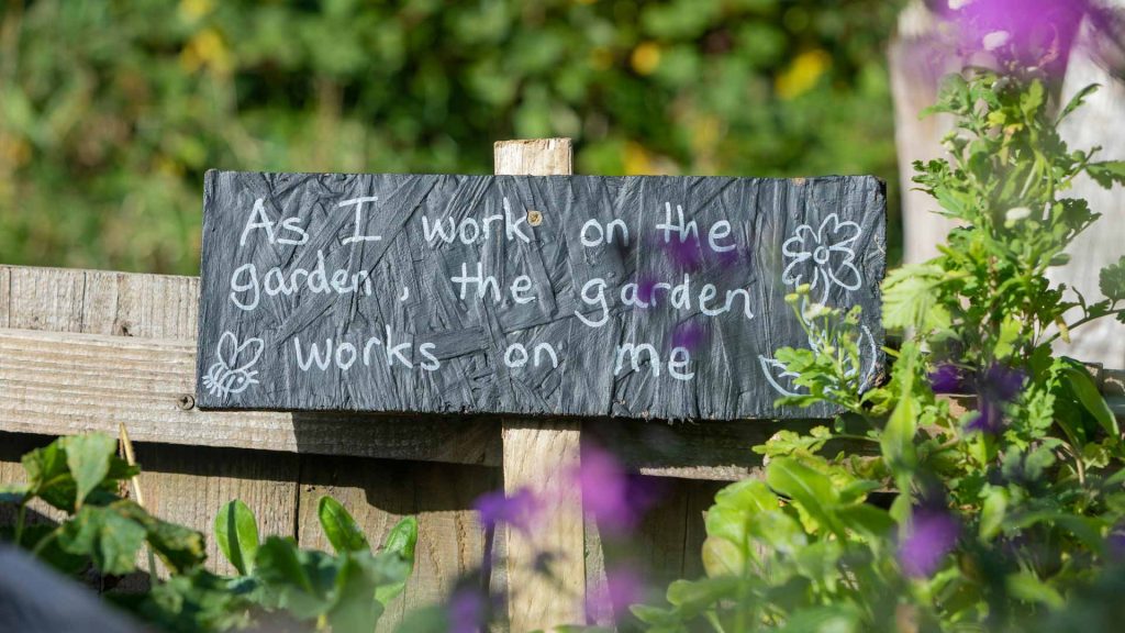 A black picket sign says, “As I work on the garden, the garden works on me.”
