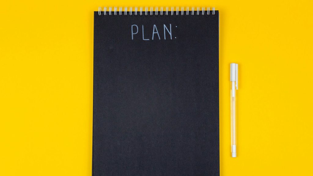 A black notepad with the word “Plan” written on it in white pen, rests on a yellow background.